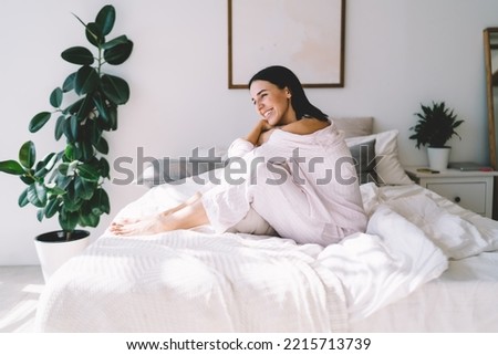 Side view of smiling young lady in nightwear sitting on bed resting on weekend in modern bedroom and looking away Royalty-Free Stock Photo #2215713739