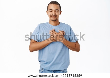 Smiling nice guy holds hands on heart, heartwarming and caring feeling, standing over white background Royalty-Free Stock Photo #2215715359
