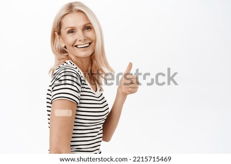 Smiling mature woman, shows thumbs up, has covid-19 vaccination band aid on shoulder, recommends people coronavirus vaccine, white background