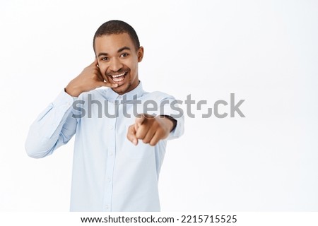 Smiling guy shows telephone gesture, points at camera, call us gesture, call center advertisement, standing over white background