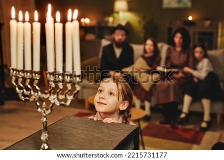 Portrait of jewish little girl looking at menorah candle in awe while celebrating Hanukkah with family, copy space