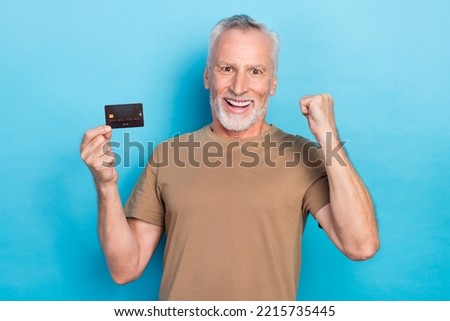 Photo of positive ecstatic pensioner gray hairstyle stubble beige t-shirt hold debit card clench fist isolated on blue color background