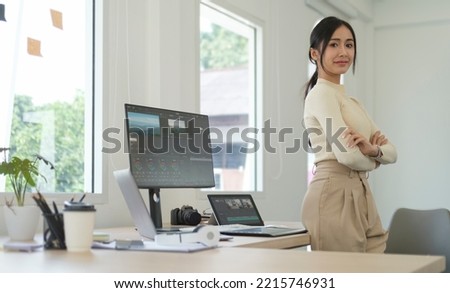 Confident female video editor  standing with crossed arms front of computer in creative office studio and looking at camera.
