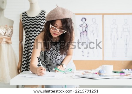 Asian woman fashion designer or dressmaker wear hat and sunglasses, drawing sketch of new collection clothes on paper, holding tape measure, working with tablet, tailoring tools on table at workplace