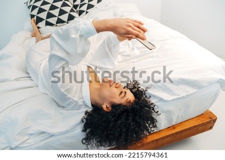 Young mixed race woman looking at cellphone browsing through social media lying upside down on the bed. Copy space.