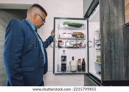 Man looking for food in an open fridge in kitchen late at night. The man looks like a hungry bachelor.