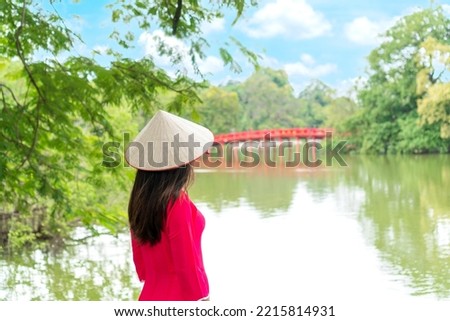 Young Asian woman tourist wearing Ao Dai (traditional Vietnamese dress) Stand by the lake, look at the Red Bridge in Hoan Kiem Lake, Hanoi, Vietnam. Copy space Royalty-Free Stock Photo #2215814931