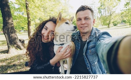 Point of view shot of cheerful married couple taking selfie with pet shiba inu dog, young people are talking, laughing and caressing the animal while looking at camera.