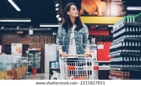 Pretty young lady in casual clothes is walking in grocery store steering shopping trolley with food inside it and looking around at shelves with products. Women and shops concept. Royalty-Free Stock Photo #2215827851