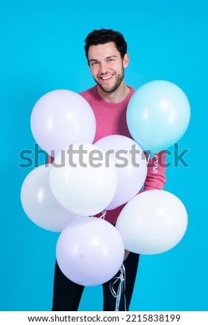 Holidays Ideas and Concepts. Smiling Funny Caucasian Guy Handsome Brunet Man With Bunch of Colorful Air Balloons in Pink Jumper Against Blue Background. Vertical Image