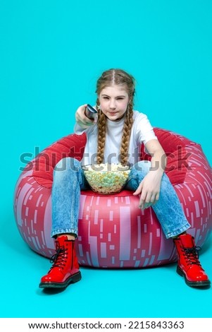 Curious Winsome Girl Teenager Girl Sitting in Chair With TV Remote Control Changing Programs Online Isolated on Turquoise Trendy Vivid Color Background.Vertical Shot