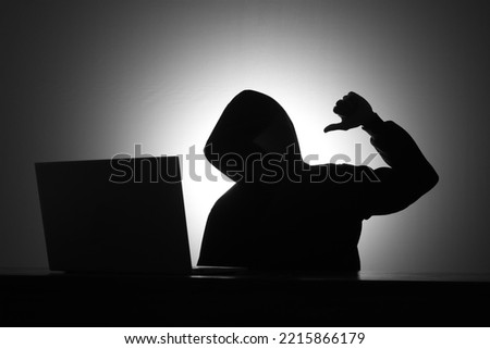 Mysterious man wearing black hoodie using laptop while mocking someone by making thumb down gesture in the dark room. Hacker and cyber security concept.