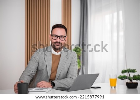 Picture of grinning young adult businessman, taking a cup of coffee, posing for the photo in his office, working.