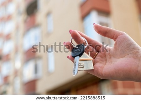 man's hand with a key and a metal key chain house hanging from the side on the background of a fence and a cottage. Construction project. moving to a new home. mortgage. renting and buying real estate