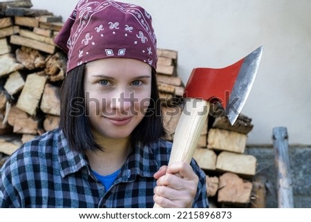 Portrait of a young adult smiling  woman stands and looks at the camera, holding an ax in front of her. On background firewood harvested prepared for cold winter, to survive the energy crisis. Royalty-Free Stock Photo #2215896873