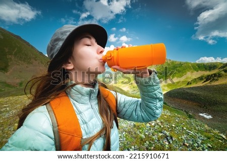 Profile photo of a young beautiful woman with long blond hair, dressed in a silver jacket, drinking from a yellow camping bottle, in the background a magnificent landscape of mountains and clouds