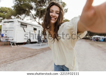 Beautiful young caucasian woman taking selfie holding out her hand to camera outdoors. Brunette wears white blouse and jeans. People emotions, lifestyle and fashion concept.