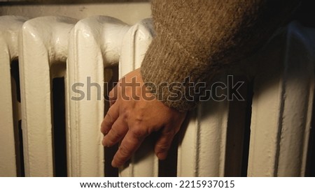 An unrecognizable hand in a warm sweater touches an old battery, tries the temperature of the battery. Heating of the house in winter. It's cold in the apartment. Rise in price of heating Royalty-Free Stock Photo #2215937015