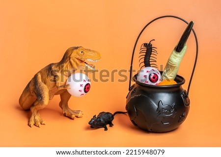 Toy dinosaur Tyrannosaurus holding eye in its paws, bowl with bat, pumpkin isolated on orange background Holiday greeting card Happy Halloween day creative minimal concept Calendar date 31 October