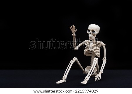 A fake human skeleton sitting on a black background and pointing with his hands at an empty space. Halloween Greeting Card