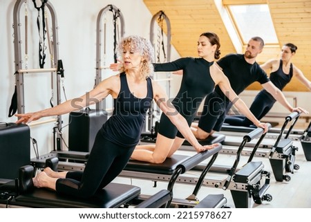 Skilled senior female trainer woman in front split on pilates reformer during class while adjusts form trains young instructors in studio. Rehabilitation, healthy lifestyle, small business concept. Royalty-Free Stock Photo #2215978627