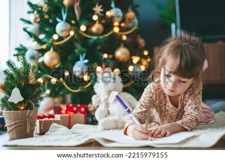 Cute little child girl writing letter to Santa Claus or writing dreams of a gift with near Christmas tree. Merry Christmas and Happy New Year!