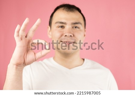 A man in a white T-shirt on a pink background holds a third molar or wisdom tooth in his hand after surgery to remove it and smiles Royalty-Free Stock Photo #2215987005