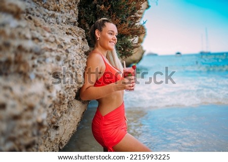 Young European woman in bikini on the beach in summer. Portrait of happy young woman smiling with a red drink. Tanned blonde girl in swimwear enjoying the beach.