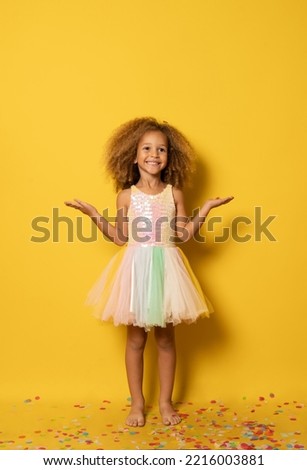 Funny little girl dressed for party in sequins and tutu blowing confetti . Isolated on yellow background.