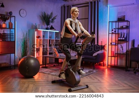 Beautiful blond caucasian sports women in sportswear cycling bike at home at night time. Cardio training, exercising legs, cardio workout indoors.