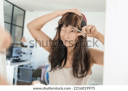 Close up of a young beautiful asian woman looking at a mirror and use a comb and hair scissors trimming bangs at home during Covid 19 pandemic lockdown. New normal, Self isolation, Social distancing. Royalty-Free Stock Photo #2216019975