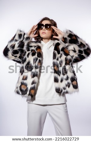 High fashion photo of a beautiful elegant young woman in a pretty leopard print fur coat, stylish sunglasses, trousers, pants posing on white background. Slim figure. Studio shot.
