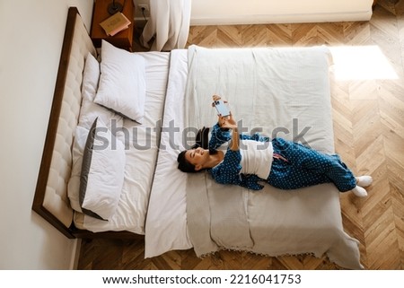 View from up on young beautiful smiling asian woman in home suit taking selfie on her phone, while lying on bed in cozy bedroom at home