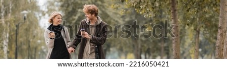 happy couple in coats holding hands and walking with paper cups in autumnal park, banner