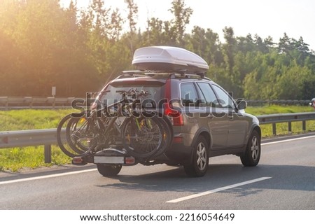 Bikes fastened on bicycle holder mounted on back side of car on country road. Brown car with roof luggage box and trunk bike rack driving on highway. Royalty-Free Stock Photo #2216054649
