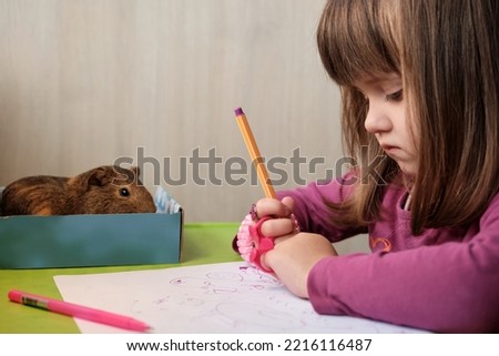 Portrait of little girl which draws at table along with a guinea pig sitting near her. Baby girl playing with his pet.