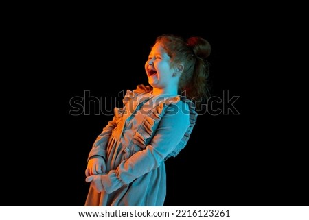 Laughing. Half-length portrait of cute cheerful little girl wearing festive dress isolated over dark background in neon light. Concept of kids emotion, facial expressions, fashion. Happy childhood