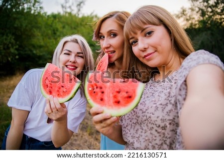 Happy young women hold slices of watermelon outdoors. Bright red berry. Juicy, sweet snack in summer on a picnic. Girls are photographed with a phone or camera. Selfie.
