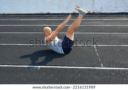 a young bald man in a gray T-shirt is doing a press on the rubber surface of the running track of the city stadium. active lifestyle, sports and physical education Royalty-Free Stock Photo #2216131989