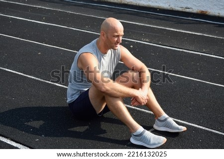 a young bald man in a gray jersey is resting after a workout on the rubber surface of the running track of the city stadium. active lifestyle, sports and physical education Royalty-Free Stock Photo #2216132025