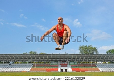 a young man in a red jersey does a jump up workout on the green lawn of the city stadium. the concept of a healthy lifestyle and sports and physical education Royalty-Free Stock Photo #2216132029