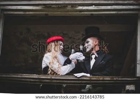 The hatter and alice in wonderland. A girl with long hair in a red hat with white makeup on her face. A man in a suit with make-up and a black hat. They drink wine. Halloween