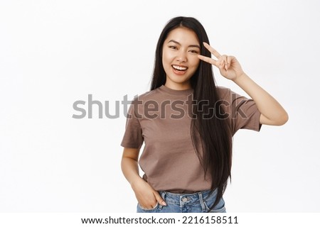 Portrait of smiling asian woman shows peace, v-sign and looks happy, shows positivity and joy, stands in tshirt over white background