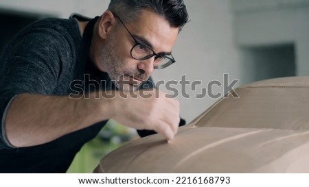 Experienced automotive designer drags across the wooden shaping tool to smooth out the surface of the car sculpture prototype. Designer with an apron works on the clay sculpture of the car. Royalty-Free Stock Photo #2216168793