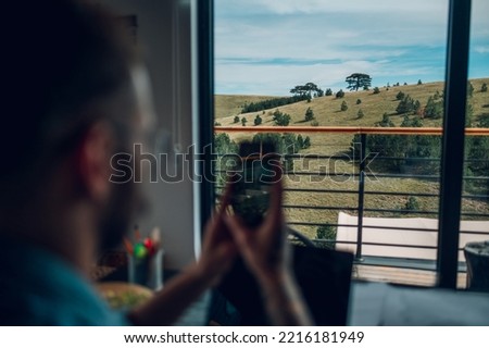 Young man working on a laptop in modern kitchen and using a smartphone. Concept of young people working from home. Focus on a window view from a cozy home.