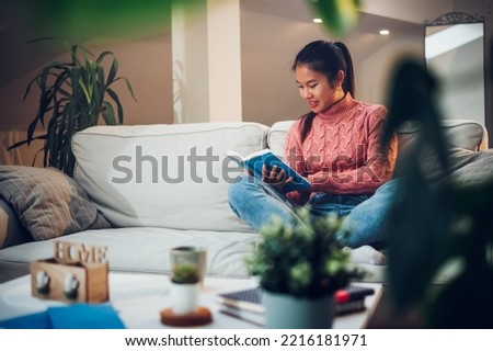 Young relaxed and happy asian woman reading a book while relaxing on sofa in cozy living room. Casual lifestyle at home. Leisure, literature and people concept. Book worm.