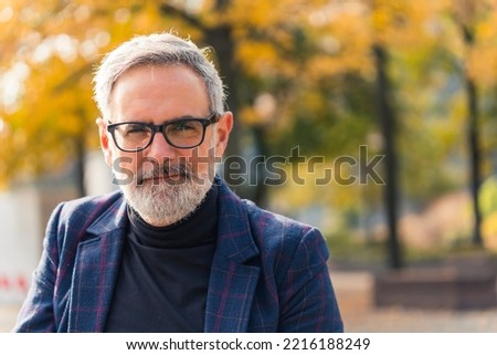 Modern confident bearded grey-haired mature man entrepreneur in a suit and having eyeglasses on, in the park with fall foliage in the background, looking at the camera. High quality photo