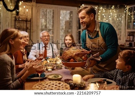 Happy man bringing roast turkey at the table during Thanksgiving dinner with his multigeneration family.
