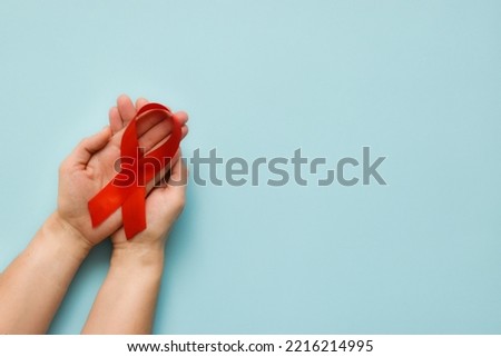 Healthcare and medicine concept - female hands holding red AIDS awareness ribbon on blue background. The symbol of The World AIDS Day or cancer or HIV Awareness Month. Top view with copy space  Royalty-Free Stock Photo #2216214995