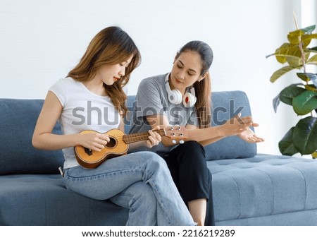 Millennial Asian young happy female lesbian LGBTQ lover couple sitting together on sofa playing small ukulele in living room.